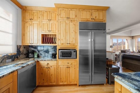 This section of the kitchen features a full-size, built-in stainless steel refrigerator/freezer, a dishwasher, a toaster oven, and a coffee maker. It has ample wood cabinetry and dark granite countertops and backsplash.