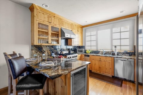 Full kitchen with stainless steel appliances (gas range, dishwasher) and granite countertops. Features natural wood cabinetry and a built-in wine cooler. A small granite bar/island with seating for two provides an eating area.