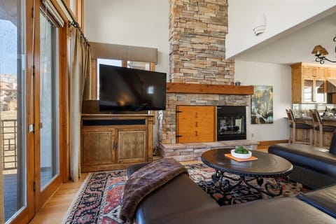 The living room features a floor-to-ceiling stone fireplace with a wooden mantel. A large flat-screen TV sits next to the fireplace. Includes a round coffee table, a sectional sofa, and large glass doors opening to an outdoor area.