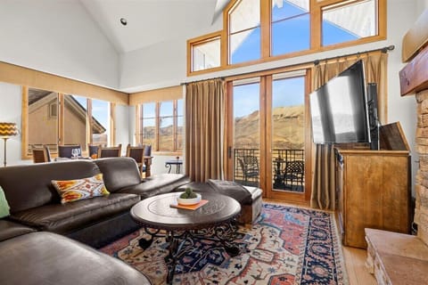 Cozy, bright living room featuring a large leather sectional sofa, flat-screen TV, and a decorative rug. Includes a balcony with stunning mountain views through large windows and glass doors. Dining area visible to the left.