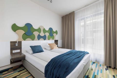A bright bedroom featuring a double bed with blue bedding, patterned carpet, and a modern headboard with colorful geometric accents.
