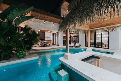 A gorgeous nighttime view from the swim-up bar, capturing the illuminated pool, lush greenery, the warm glow of the rancho’s teak ceiling, and the house lit beautifully in the background.