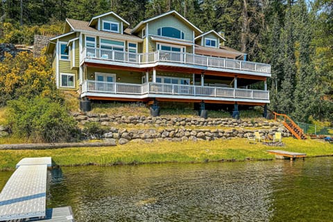 Bright lakeside exterior — dual wraparound decks, stone retaining wall, private lawn, dock ramp, and tall pines. Every level faces the water