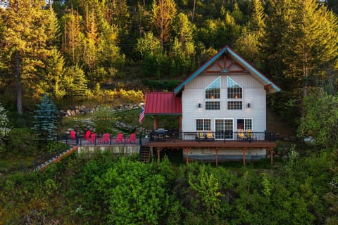Twilight aerial of A-frame cabin with soaring gable windows, wraparound deck, red Adirondack chairs & string lights nestled in forest — the shot you screenshot before booking