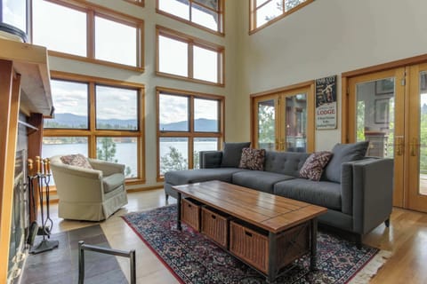 Sunlit great room - soaring two-story windows, sectional sofa, wood-plank coffee table, fireplace. The lake view stops you in your tracks the moment you walk in