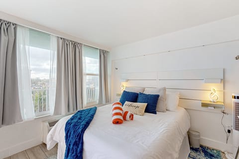 Sun-filled beach stay bedroom featuring a plush queen bed, crisp linens, and coastal-blue accents. Large windows invite natural light, creating a welcoming and relaxing oceanside escape.
