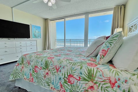 Wake up to sweeping ocean views in this bright, coastal primary bedroom featuring a plush king bed, beach-inspired décor, flat-screen TV, and private balcony access—perfect for a relaxing beachfront stay.