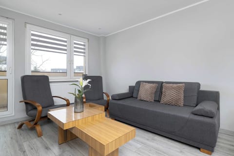 A bright living room with a gray sofa, matching armchairs, and a wooden coffee table. Soft natural light enters through large windows, creating a cozy atmosphere. The neutral décor makes the space feel clean and modern.
