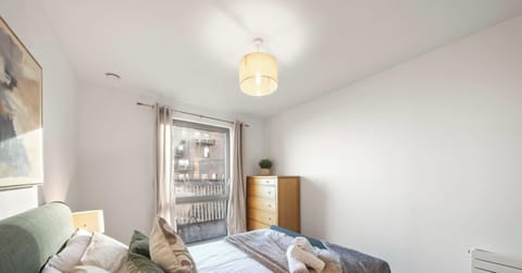 A bright and modern bedroom features a neatly made bed with sage green and patterned accent pillows. To the side, a tall wooden dresser stands near a large window that fills the clean, white-walled space with natural light.