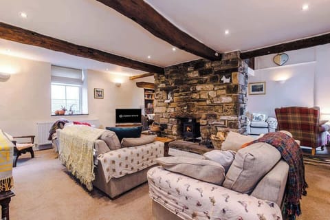 A cozy living room features a large rustic stone fireplace with a wood-burning stove, surrounded by comfortable sofas and warm wooden beams across the ceiling.