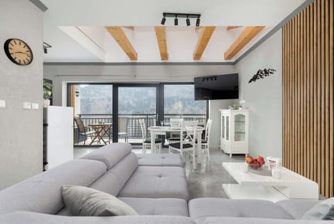 A bright living room with a large sectional sofa, wooden ceiling beams, and floor-to-ceiling glass doors leading outside. Natural light fills the space, highlighting a clean and modern design.