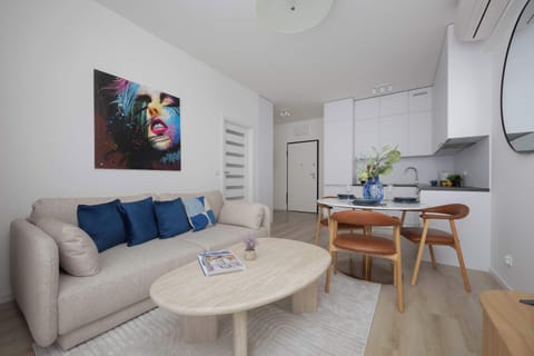 A cozy living room with a beige sofa and a small round coffee table. A dining table with chairs is visible in the background near the window.