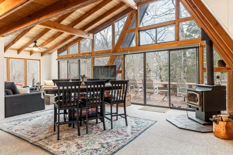 The vaulted great room combines dining and living areas beside a freestanding wood stove and a wall of windows opening toward the deck.