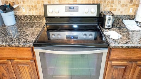 This kitchen boasts a modern Whirlpool glass-top electric range with spacious oven, perfect for home-cooked meals. Set against granite counters & wood cabinetry, it includes essentials like a toaster and utensils for convenient, ready-to-use cooking.