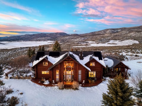 Exterior of a large log cabin home at sunset, covered in snow, with a mountain view.