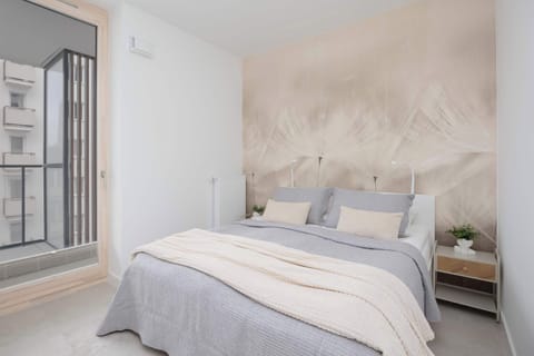 A bright bedroom with a double bed placed near a large window. Soft neutral bedding and light walls create a calm atmosphere. Natural light fills the space.
