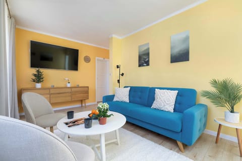 A bright living room with yellow walls, a blue sofa, and a white coffee table. Decorative pillows add color and contrast. Natural light enters through large windows.
