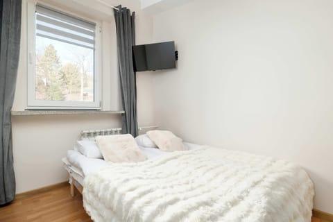 A brightliving area with a neatly made double bed, light-colored walls, wooden flooring, and a window bringing in natural daylight.