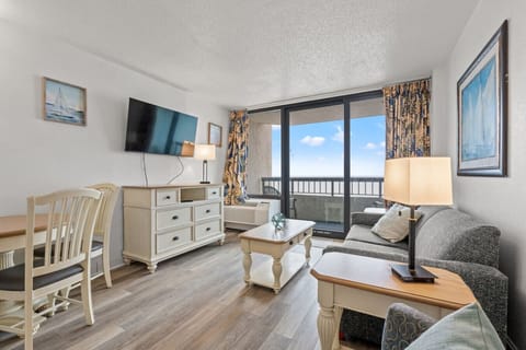 Bright oceanfront living room with floor-to-ceiling windows, natural light, sofa, chair, sleeper sofa, queen Murphy bed, Smart TV, and dining area. Private balcony showcases stunning shoreline views.