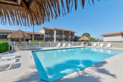 Another ground-level view of the Sea Sands pool area with palapa umbrellas, loungers, and neighboring condo building. Highlights a sunny spot for relaxing between beach trips.