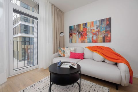 A cozy living room with a light-colored sofa, orange throw blanket, and round coffee table. Large windows with sheer curtains let in natural light. A colorful abstract painting decorates the wall.