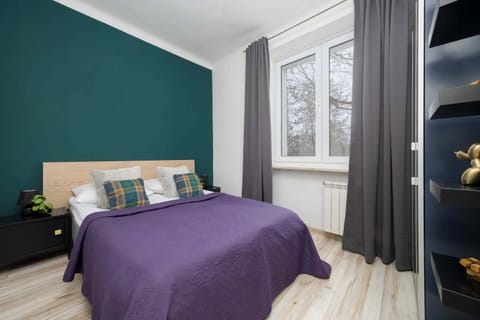 A bedroom with a deep green accent wall and a bed covered with a purple duvet. Soft curtains frame the window and let in natural light. The space feels cozy and calm.