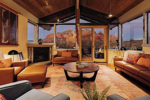 Unwind in your cozy living room with panoramic red rock views through floor-to-ceiling windows, complete with fireplace and rustic beams. A TV is hidden within the cabinetry just left of the fireplace. 