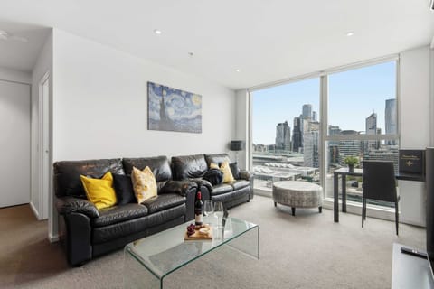 Living room with black leather sofas, glass coffee table and floor‑to‑ceiling windows offering city skyline views.