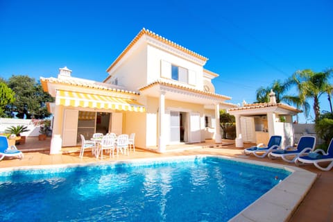 Stunning exterior view of Casa Destino, a professionally cleaned 3-bedroom villa featuring a large private swimming pool, sun loungers, outdoor dining furniture and a shaded terrace with striped awning, set against the backdrop of the beautiful Algarve countryside near Lagos, Portugal