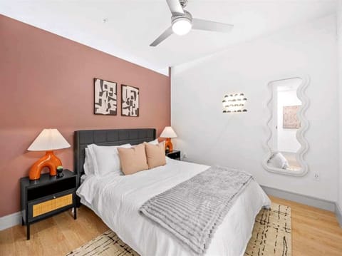 This cozy bedroom features a soft, inviting queen-sized bed with plush pillows, set against a warm terracotta accent wall. Modern lamps add a touch of elegance, while a ceiling fan ensures comfort. The minimalist decor and light wood floors create a 