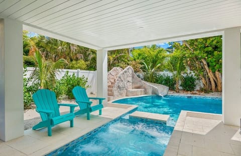 Resort-style backyard featuring a sparkling private pool, shallow lounging ledge, and covered patio seating. Surrounded by lush tropical greenery, this inviting outdoor space is perfect for relaxing, swimming, and enjoying sunny coastal days.