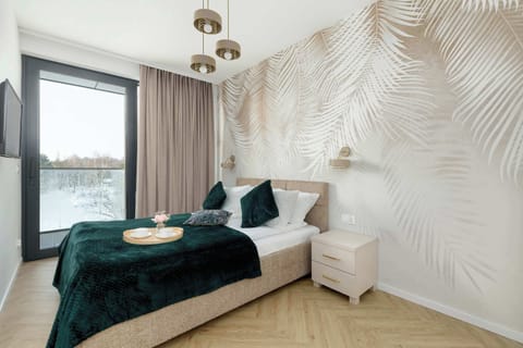 A bedroom featuring a green bedspread and decorative wall panels behind the bed. Natural light enters through a large window. The room feels calm and tastefully designed.
