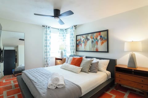 A stylish bedroom featuring a large Roku TV with soundbar, ceiling fan, dual nightstands, standing mirror, and a relaxed vibe ideal for movie nights.