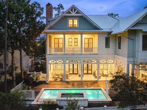 Charming coastal cottage with glowing windows, wraparound porches, and sparkling pool creating perfect evening ambiance.
