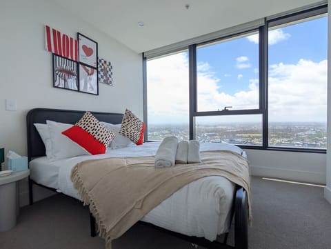 High-floor bedroom with sweeping panoramic views stretching across the entire city to the horizon. Bold pop-art wall prints add personality, while the clean white bedding and corner windows keep the space feeling open and light-filled.