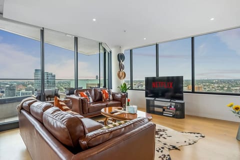 Expansive corner living room with wraparound floor-to-ceiling windows, leather sofas, a cowhide rug, and a large smart TV with Netflix. The 270-degree city and bay views make this a genuinely impressive space to spend time in.