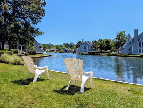 4006 Brighton Trail - view - Picturesque waterfront view featuring two white Adirondack chairs on a grassy lawn directly overlooking the serene community lake, with townhomes visible along the sunny opposite shore.