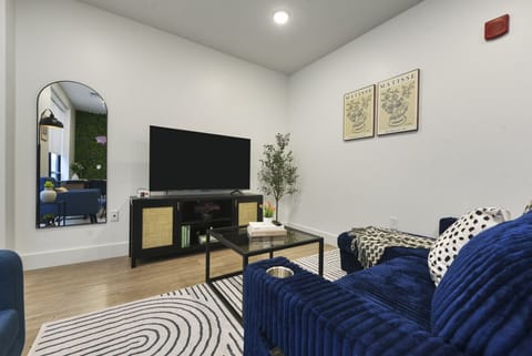 Modern living room with luxurious blue velvet sofa, large flat-screen TV, arched mirror, and vibrant green living wall – perfect for stylish and comfortable stays.