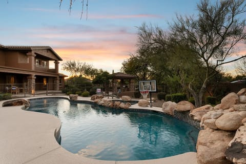 Stunning desert resort-style pool with natural rock accents surrounded by mature landscaping and mountain views at sunset.