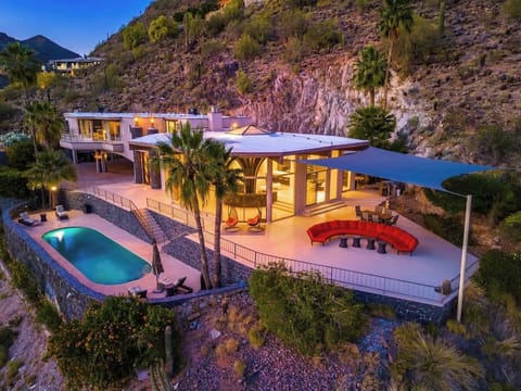 A striking desert retreat nestled into dramatic rocky hillsides, featuring modern architecture with illuminated interiors, a private pool, terraced outdoor spaces, and lush desert landscaping.