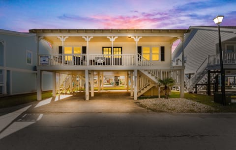 A charming beach house welcomes guests with its colorful sunset backdrop and elevated coastal design featuring covered parking below.
