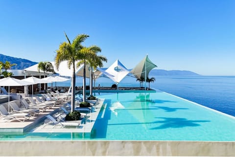 Gorgeous rooftop infinity pool overlooking the ocean, lined with sun loungers and palm trees. Enjoy the perfect blend of luxury and tranquility with uninterrupted views of the bay and mountains.