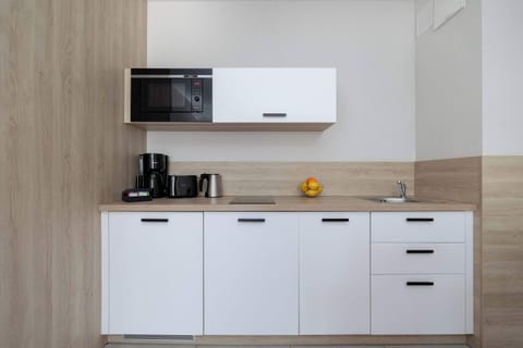 A sleek kitchenette with white cabinets, a microwave, coffee machine, kettle, and wood-accented backsplash.