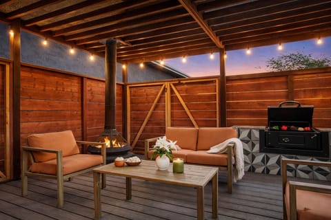 A cozy, covered outdoor lounge featuring wooden paneling, string lights, a BBQ grill, and an outdoor fireplace for evening gatherings.