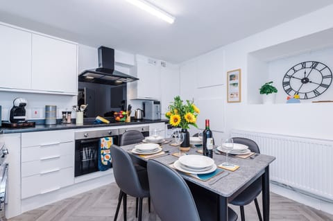 A modern fitted kitchen with sleek white units, contrasting worktops, and integrated oven, hob with extractor, and washing machine. A dining table with seating sits centrally, complemented by bright lighting and contemporary finishes.