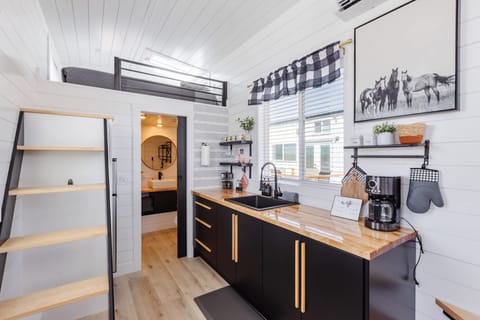 Bright modern tiny home kitchen with loft sleeping area, open shelving, coffee station, and stylish finishes throughout. Spacious layout with natural light and smart storage creates a cozy, functional space for relaxing stays.