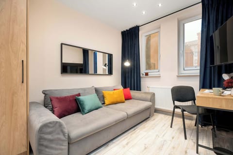 A compact living room with a gray sofa and colorful accent pillows. A small desk and chair are placed near the window. Dark curtains contrast with the light flooring.