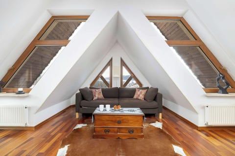 A bright attic living area with a sloped wooden ceiling and skylight windows. A gray sofa and wooden coffee table sit on polished wooden floors, creating a warm, modern feel.