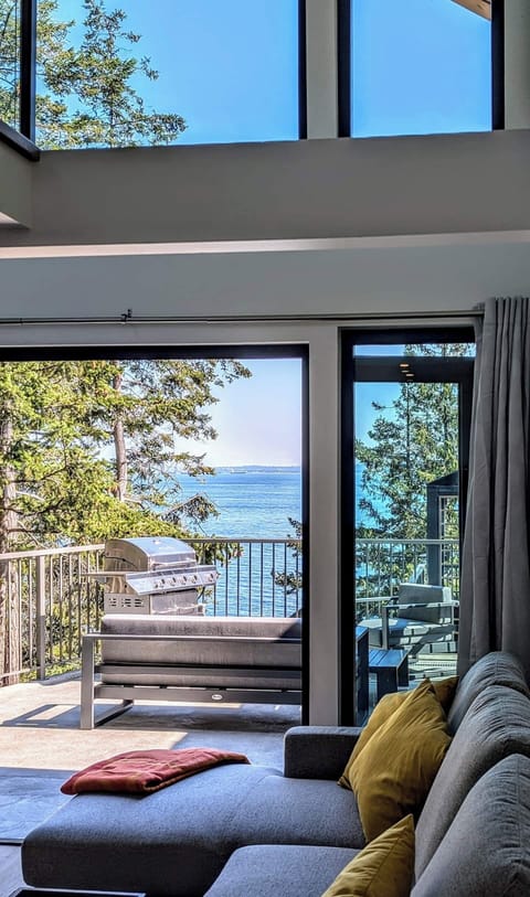 Bright ocean-view living room with floor-to-ceiling glass, cozy seating, and direct access to a private patio with BBQ, ideal for sunsets, sea air, and relaxing.