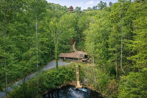 Aerial view of a secluded mountain retreat nestled among trees, featuring a rustic home with pond access via wooden dock.